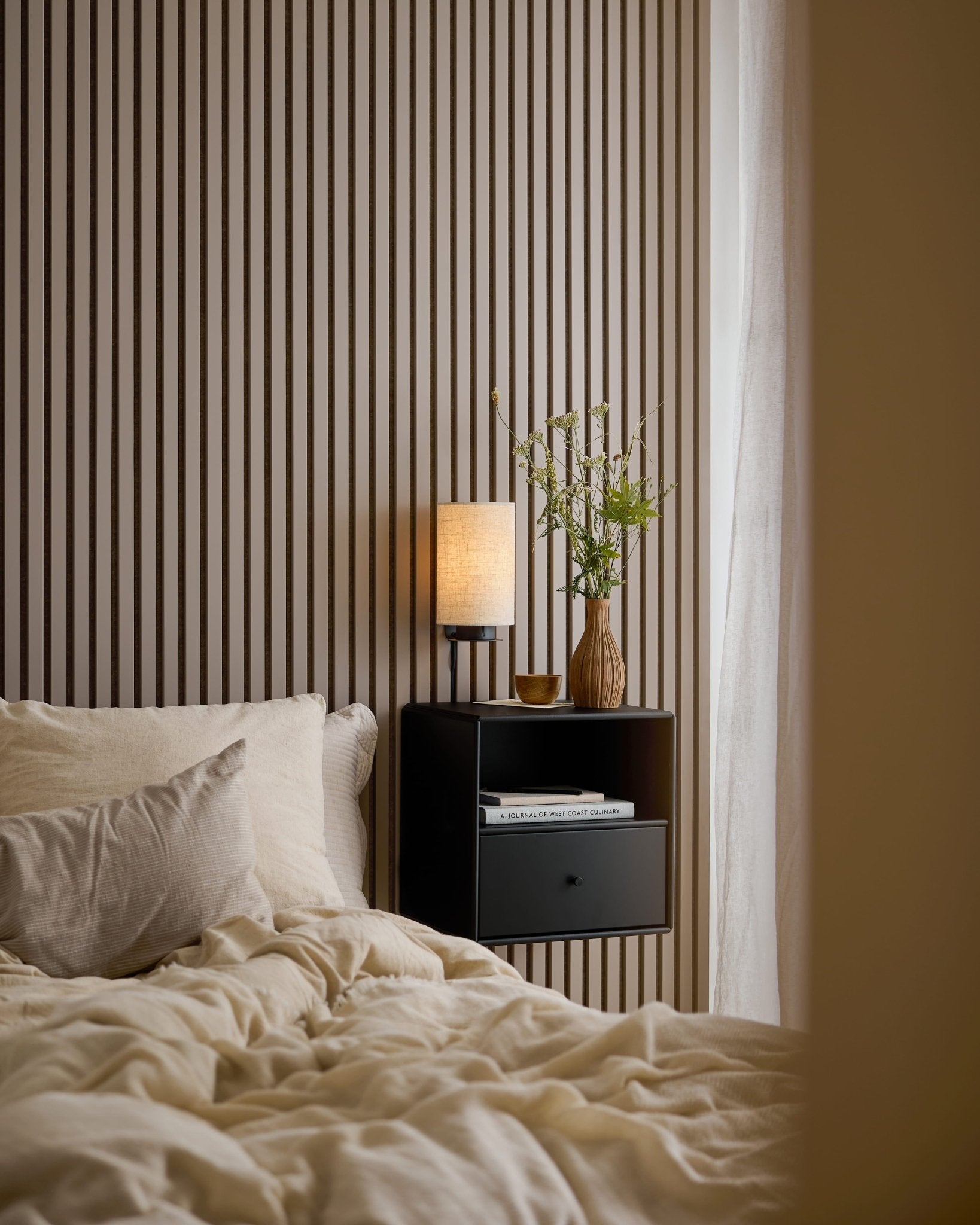 Ein Bett mit cremefarbener Bettwäsche, Büchern und einer Lampe auf einem schwarzen Nachttisch neben einer WoodUpp Akupanel Dark Cashmere-Akustikplatte, die für Stil und Schalldämpfung sorgt.